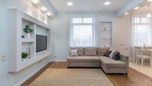 A modern living room with a sectional sofa, a flat-screen TV mounted on the wall, a dining area with a table and chairs, and a large window with sheer curtains. - Lux apartment with view on Svisloch (Minsk)