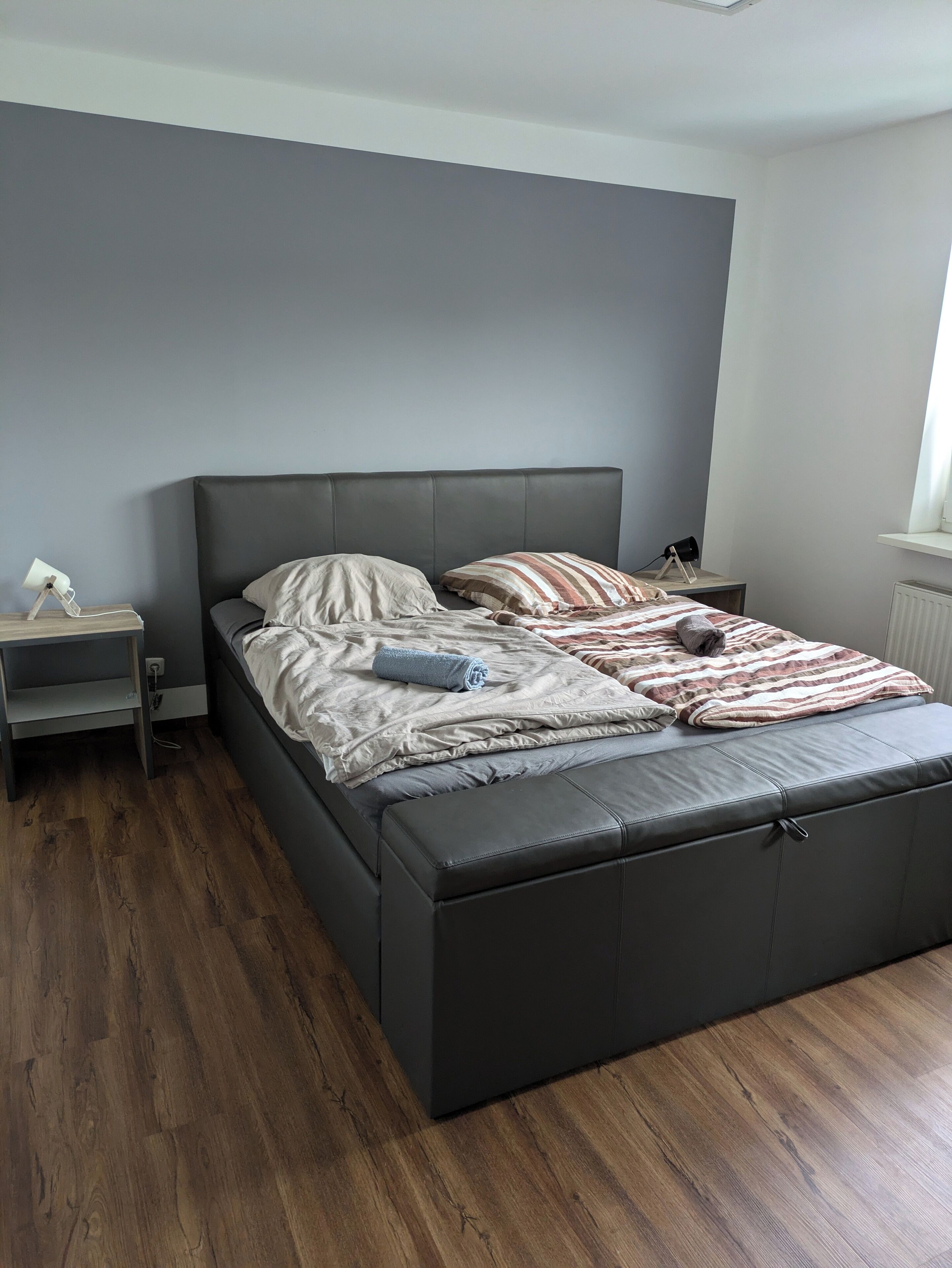 Ein modernes Schlafzimmer mit einem grau gepolsterten Bett, einem Holfnachtschränkchen und einem Nachttisch.