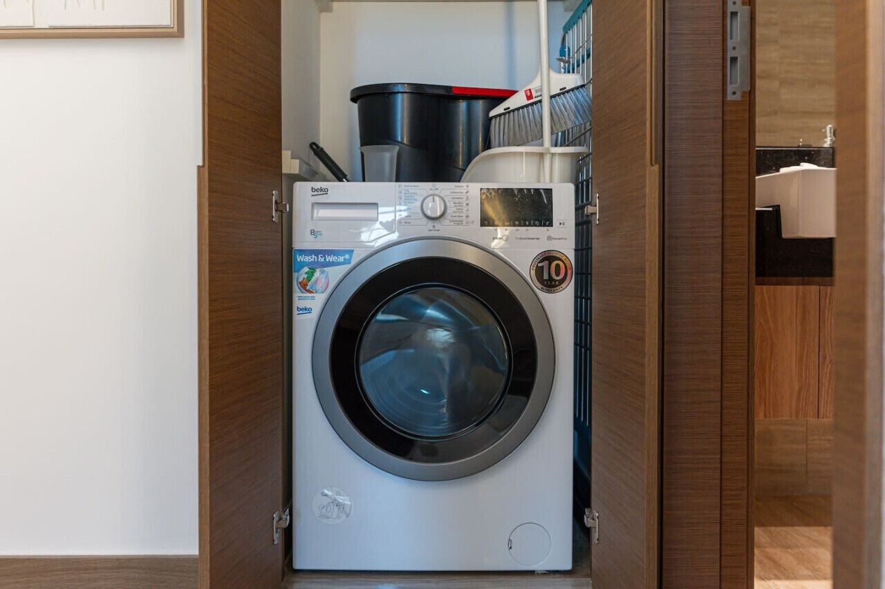 Una lavadora de carga frontal en un cuarto de lavado con puerta de madera, un estante superior que contiene productos de limpieza y una ventana pequeña que muestra un baño con un lavamanos.