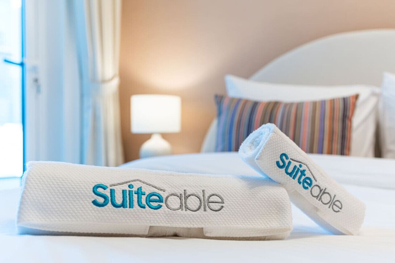 Een net opgemaakt bed met een witte dekbedovertrek en twee opgerolde handdoeken met het logo 'Suiteable'.