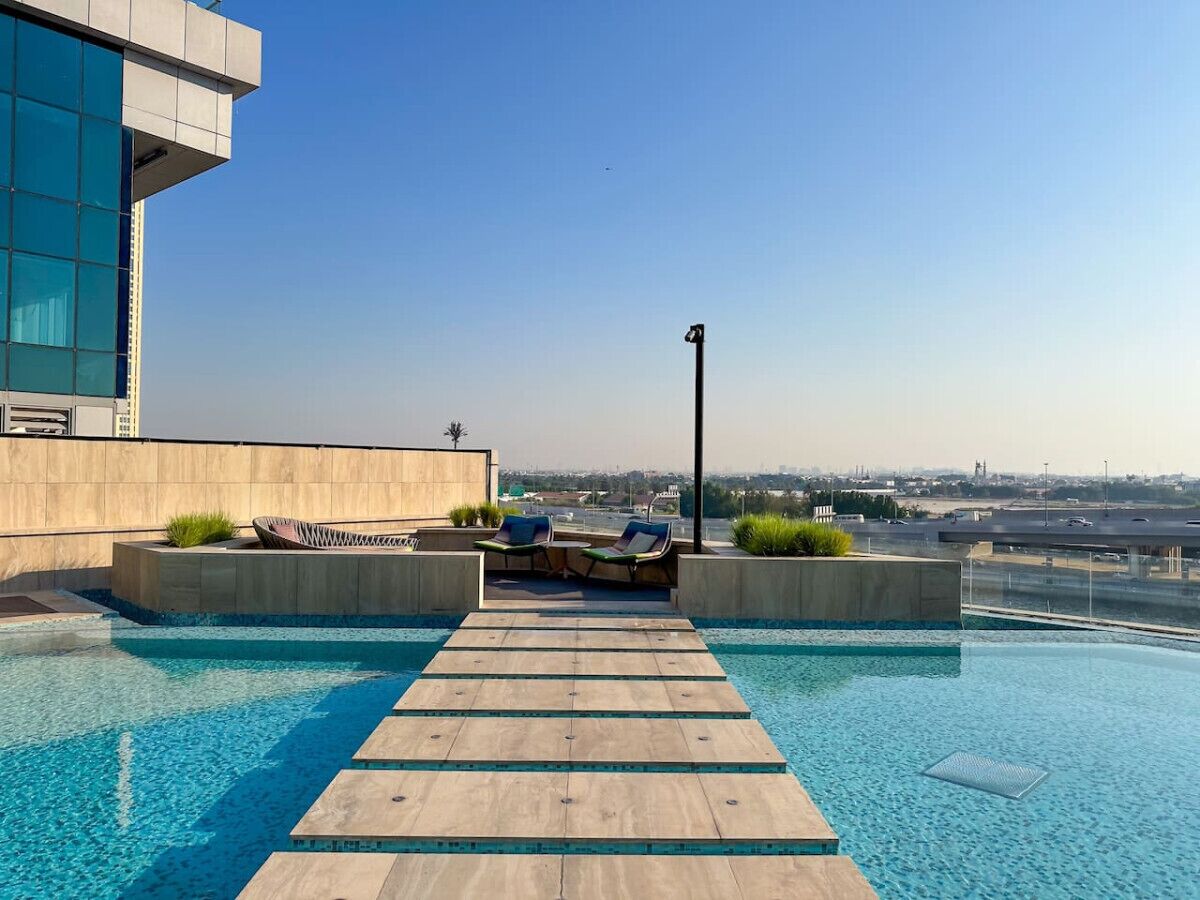 Une piscine à débordement située sur le toit, avec une terrasse en bois et des chaises longues, entourée d’une architecture moderne et offrant une vue dégagée sur le panorama urbain.