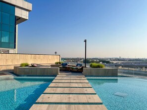 A rooftop infinity pool with wooden deck and lounge chairs, surrounded by modern architecture and a clear view of the city skyline.