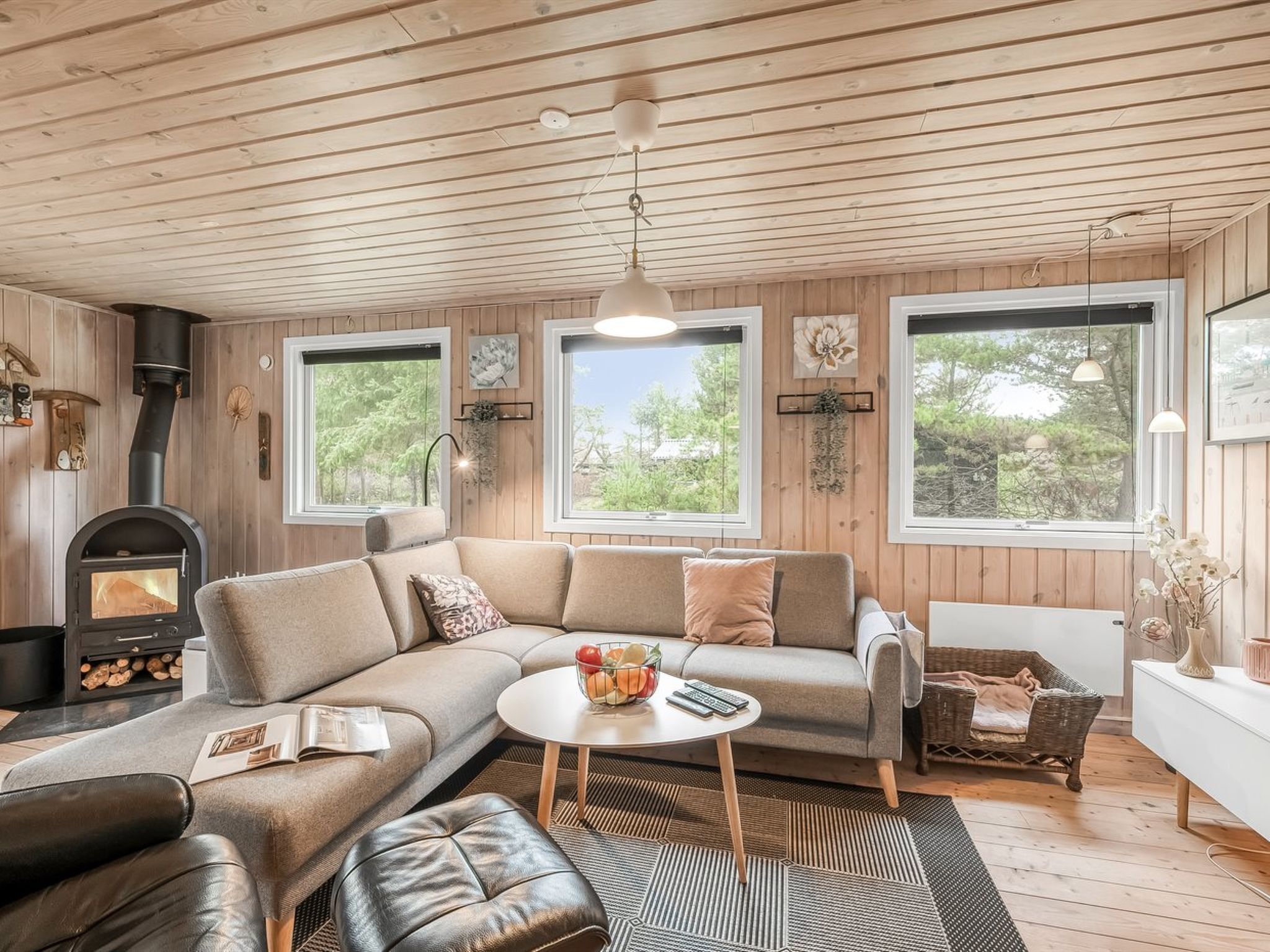A cozy living room with a sofa, ottoman, and coffee table. A fireplace is visible on the left, and there are large windows providing natural light.