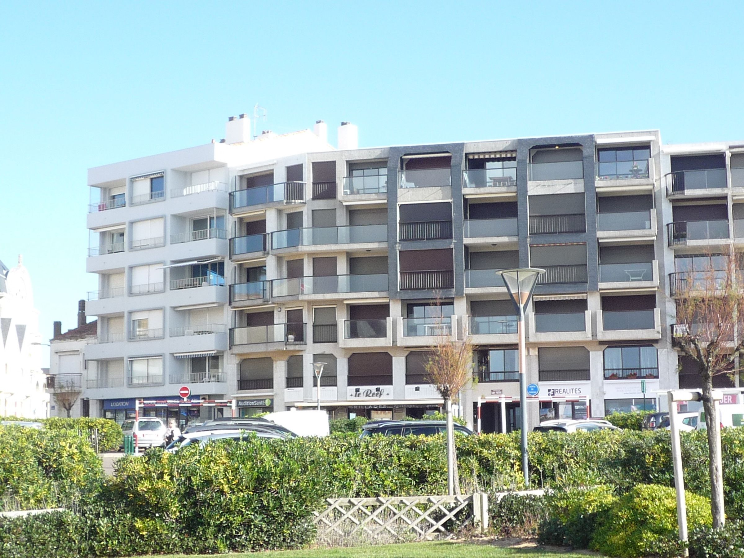 A multi-story residential building with balconies, a parking area, and a street in front.