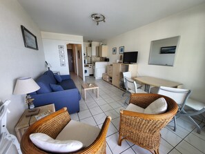A compact living space with a sofa, two wicker chairs, a small table, a TV, and a kitchenette.