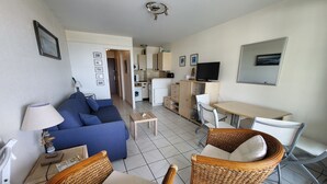 Un espacio habitable compacto con un sofá, dos sillones de mimbre, una mesita, televisor y una kitchenette.