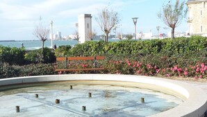 A waterfront promenade with a circular water feature, benches, and a view of the sea and distant buildings.