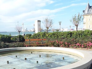 A waterfront promenade with a circular water feature, benches, and a view of the sea and distant buildings.