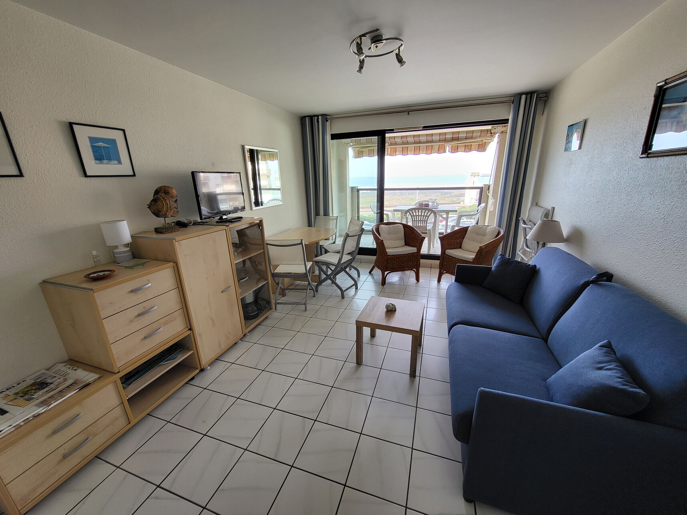 A compact living room with a blue sofa, a wooden cabinet, a small table, and a balcony with chairs.