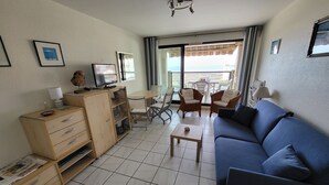A compact living room with a blue sofa, a wooden cabinet, a small table, and a balcony with chairs.
