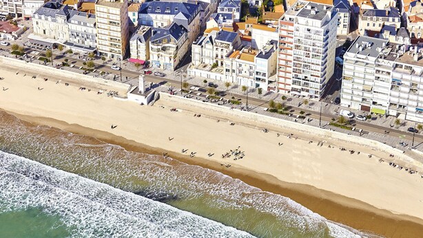 Vista aérea de una zona costera con edificios a lo largo de la playa y olas rompiendo en la orilla.