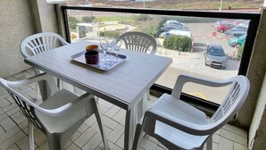 Un balcón con una mesa y sillas, una bandeja con una taza y vasos, y vista al océano y al estacionamiento.