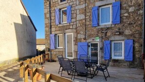 Un edificio de piedra con contraventanas azules, una terraza de madera con asientos exteriores y un cielo despejado.