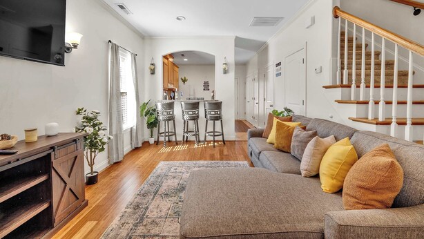 A living room with a sofa, a TV, a wooden floor, a staircase, and a kitchen in the background.