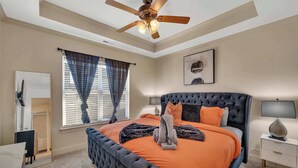 A bedroom with a tufted headboard, an orange bedspread, a ceiling fan, a window with curtains, and a bedside table with a lamp.