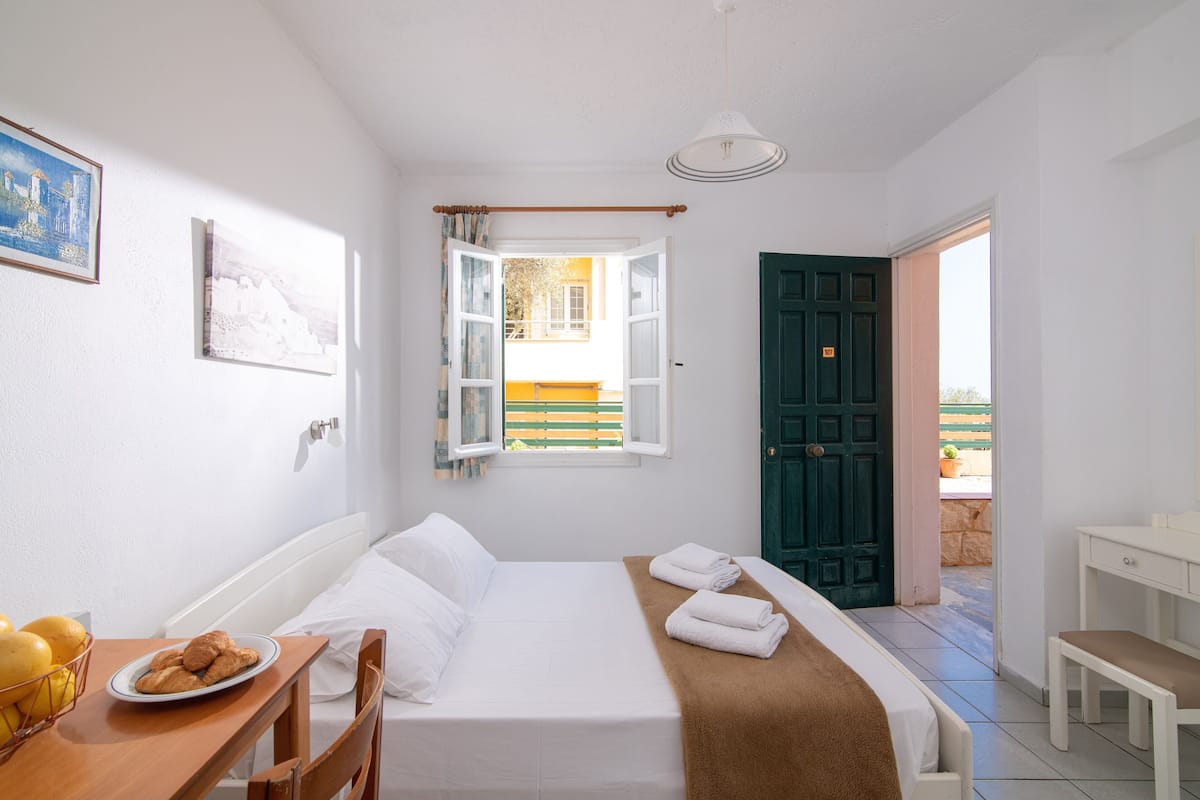 Un dormitorio ordenado con una cama, una mesita de noche con un plato de galletas, una ventana con cortinas y vistas al exterior.
