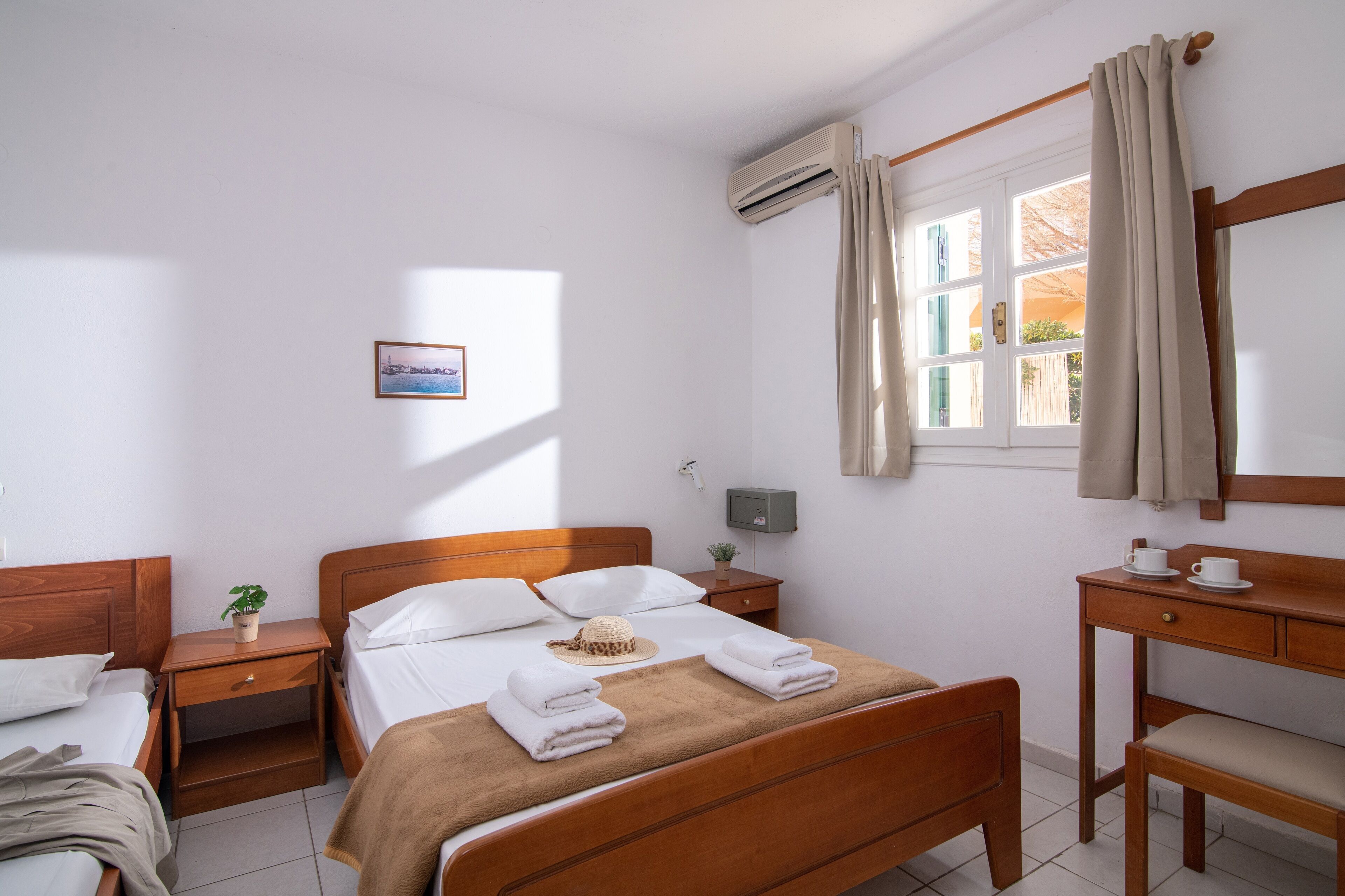 Habitación de hotel con cama de madera, un escritorio, una silla, una mesita de noche, una planta en maceta y una ventana con cortinas.