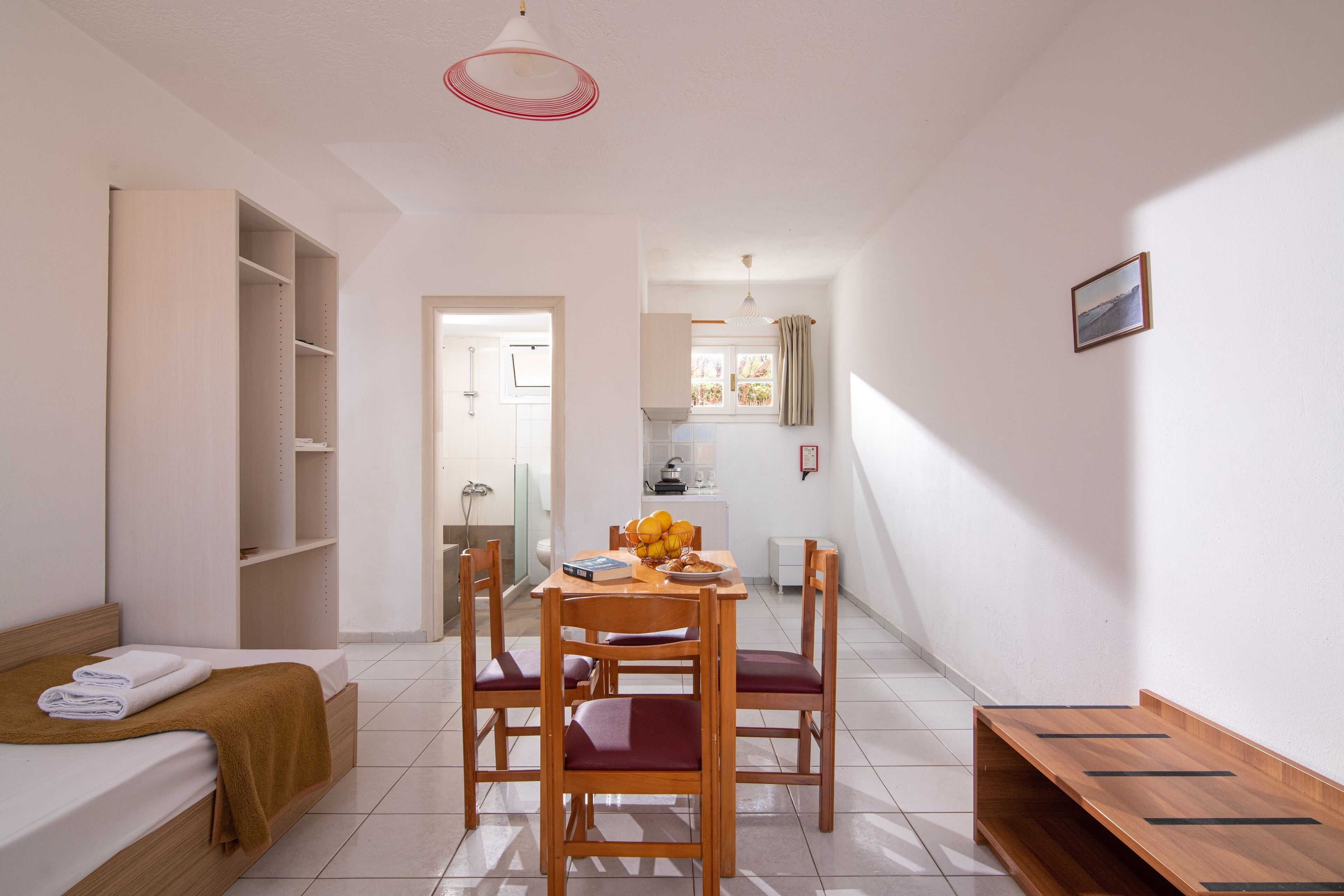 A small, well-lit room with a bed, a dining table with chairs, a wooden bench, and a kitchen area in the background.