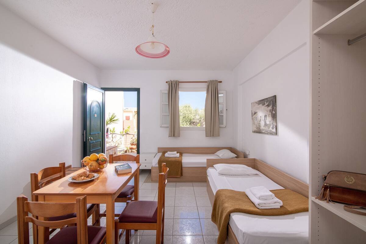 Habitación de hotel compacta con zona de comedor, cama con manta marrón, pequeña mesa con sillas y ventana con cortinas.