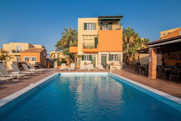 A swimming pool with lounge chairs and a building with balconies. - Golden Valentin Apartments (Hersonissos)