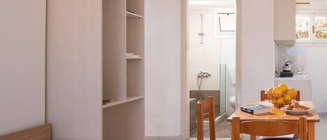 A compact hotel room with a bed, a wooden chair, a small table with a fruit bowl, and a kitchenette.