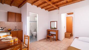 Una habitación compacta con techo de madera, una zona de comedor con mesa y sillas, un pequeño escritorio de madera con espejo, una cama y un baño visible a través de una puerta abierta.