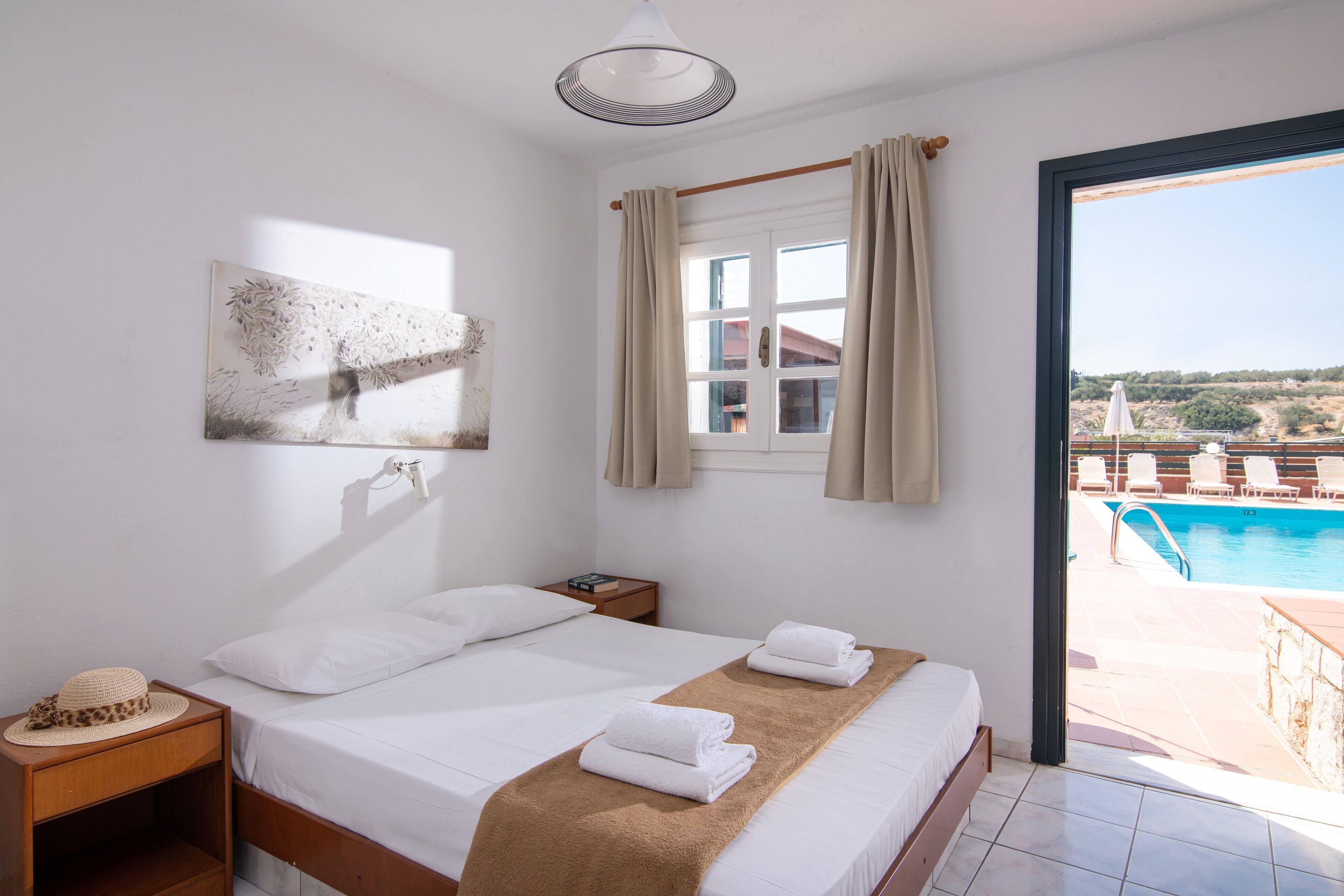 A hotel room with a bed, a nightstand, a painting, a window with curtains, and a view of a swimming pool.