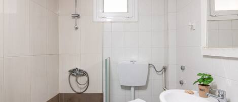 A bathroom with white tiles, a glass shower enclosure, a toilet, a sink with a plant, and a window above the toilet.