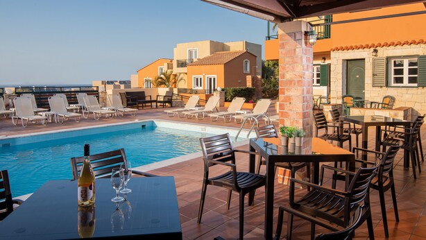 A poolside area with lounge chairs and tables, a pool, and buildings in the background. - Golden Valentin Apartments (Hersonissos)