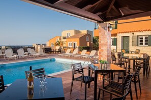 A poolside area with lounge chairs and tables, a pool, and buildings in the background. - Golden Valentin Apartments (Hersonissos)