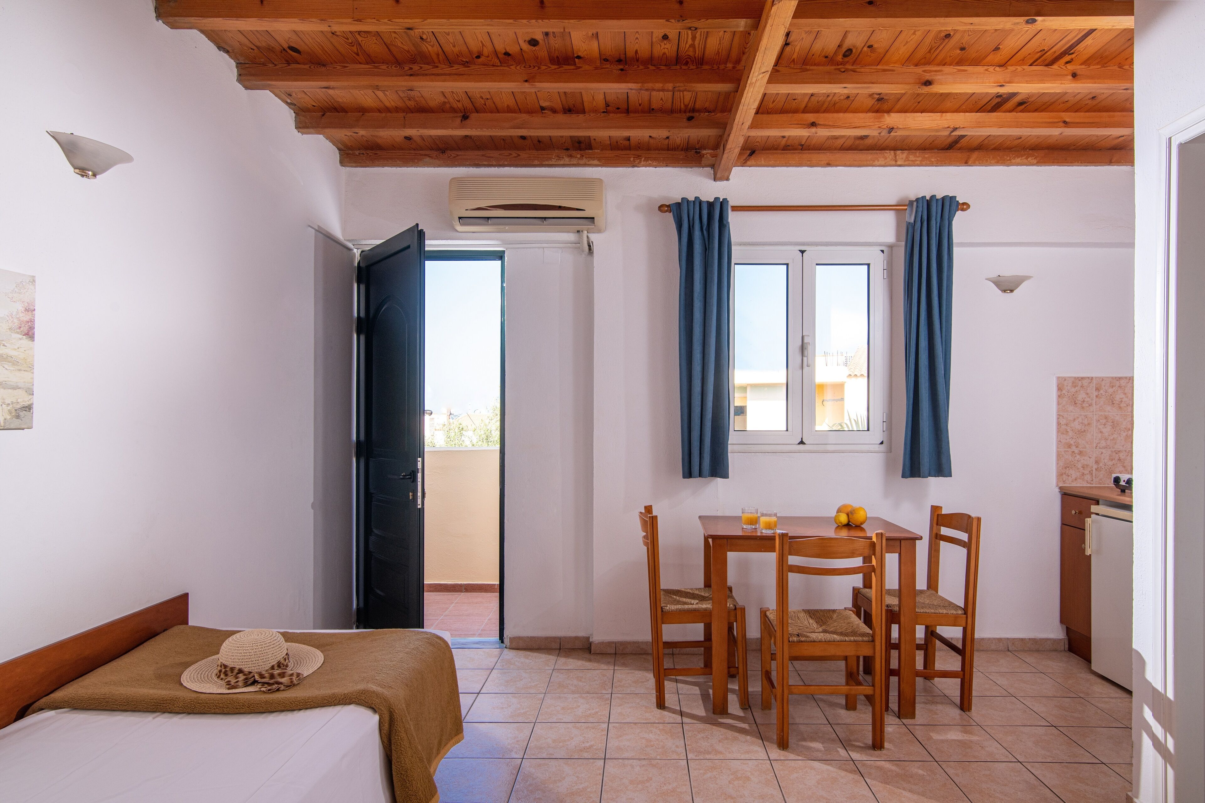 A room with a bed, a table with chairs, a window with curtains, an air conditioning unit, and a view of the outside.