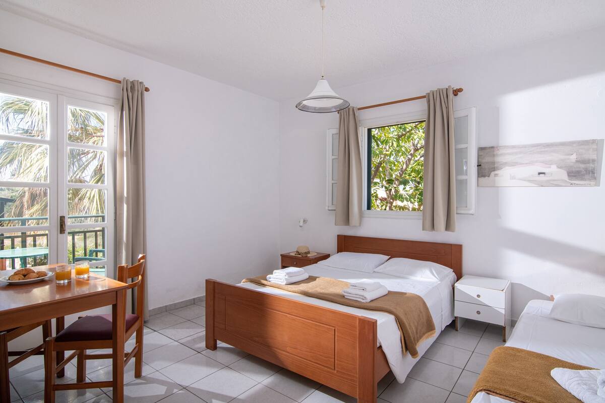 Un dormitorio con una cama de madera, una mesita de noche, cortinas en la ventana y vistas al exterior con vegetación.