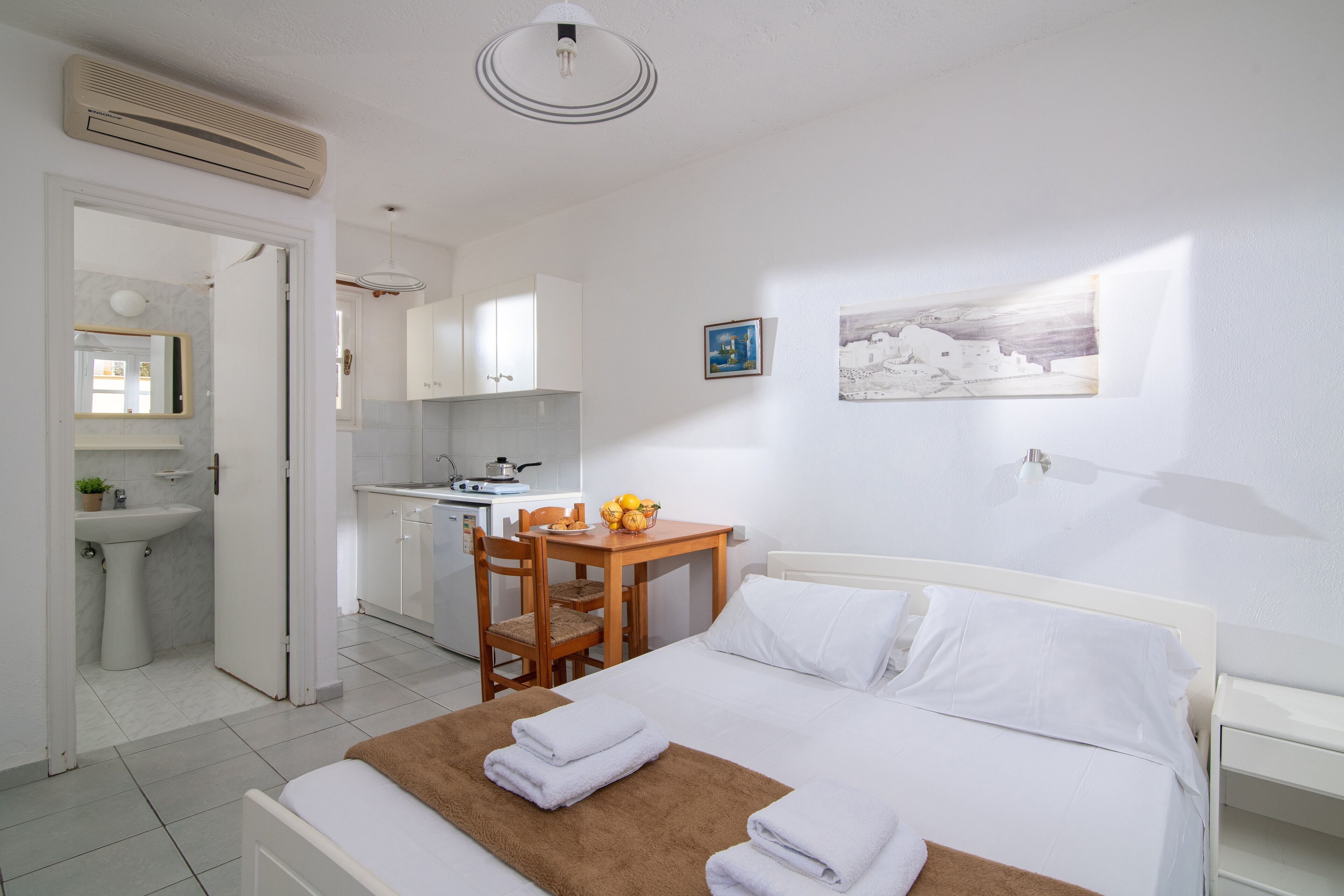 Habitación de hotel con cama, una mesita con frutero, una silla, escritorio con computadora y una cocina visible a través de una puerta abierta.