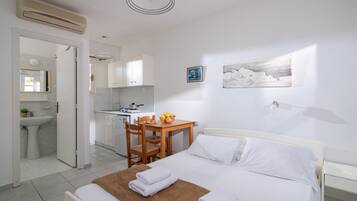 Habitación de hotel con cama, una mesita con frutero, una silla, escritorio con computadora y una cocina visible a través de una puerta abierta.