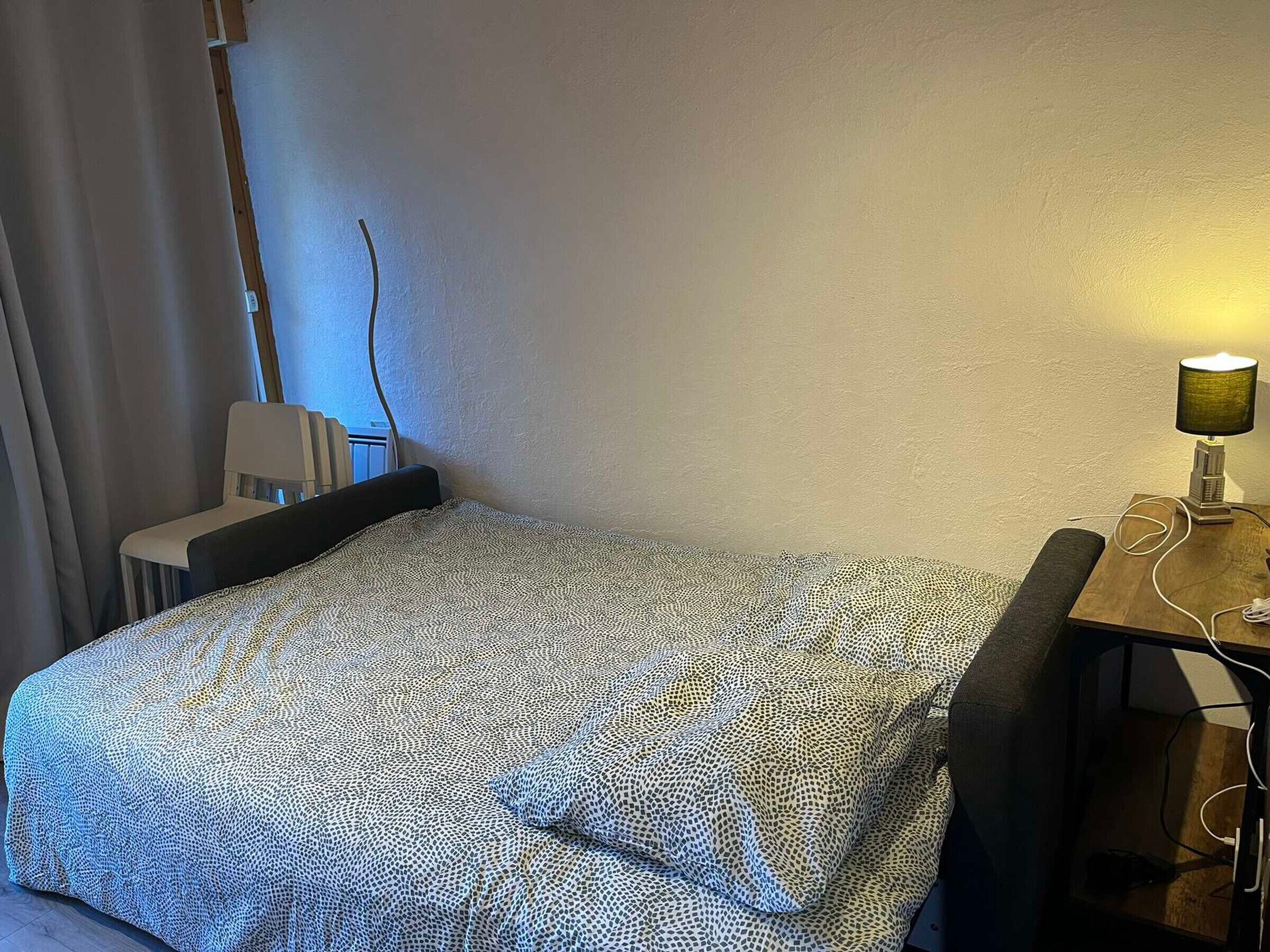 A small, neatly made bed with a patterned bedspread, a bedside table with a lamp, and a chair against the wall.
