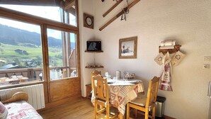 A cozy room with a wooden table and chairs, a bed with a quilt, a window with a view of the countryside, a clock, and a television on the wall. - Alcove studio for 3 with terrace near slopes and parking in Praz-sur-Arly (Praz-sur-Arly)