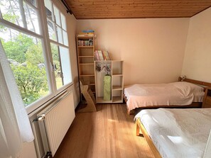 Une chambre avec deux lits, un plancher en bois, un radiateur, une fenêtre donnant sur des arbres et une bibliothèque contenant des livres et un sac vert.