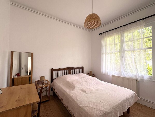 A bedroom with a wooden bed, a wooden desk, a chair, a mirror, a hanging light fixture, and a window with sheer curtains. - Charming villa with garden, 100m from Arcachon Bay, near beaches and shops, Wi-Fi (Lège-Cap-Ferret)