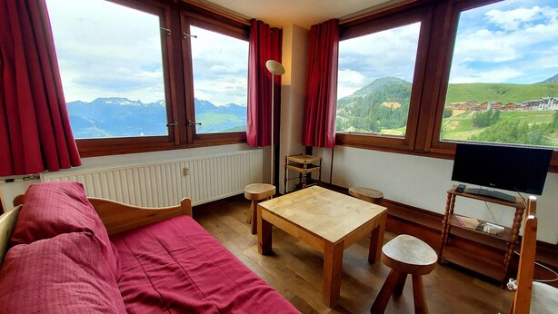 A cozy room with a wooden floor, a red sofa, a wooden table, a small stool, and a television on a wooden stand. Large windows offer a view of mountains and a village. - Comfortable apartment, close to slopes, pets allowed (La Plagne-Tarentaise)