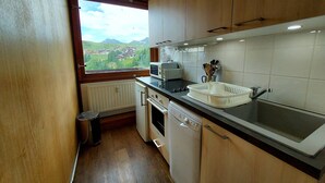 A compact kitchen with wooden cabinets, a built-in microwave, a sink, and a window offering a view of a town and mountains. - Comfortable apartment, close to slopes, pets allowed (La Plagne-Tarentaise)