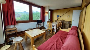 A cozy room with a wooden sofa, a dining table, and chairs. There is a TV on a small stand, a lamp, and a window with curtains. - Comfortable apartment, close to slopes, pets allowed (La Plagne-Tarentaise)