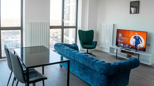 A modern living room with a glass table, blue sofa, green armchair, and a TV displaying a show. - Luxury two Bedroom Apartment (Barking)