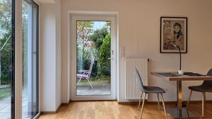 A modern room with a wooden floor, a dining table with chairs, a radiator, and a door leading to an outdoor area with a chair and greenery.