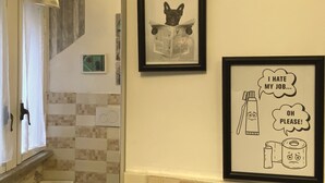 A bathroom with a tiled backsplash, a framed picture of a dog reading a newspaper, and another framed picture with a toilet paper roll and a toothbrush expressing dislike for their job. - Navona "The Corner"  (Roma)