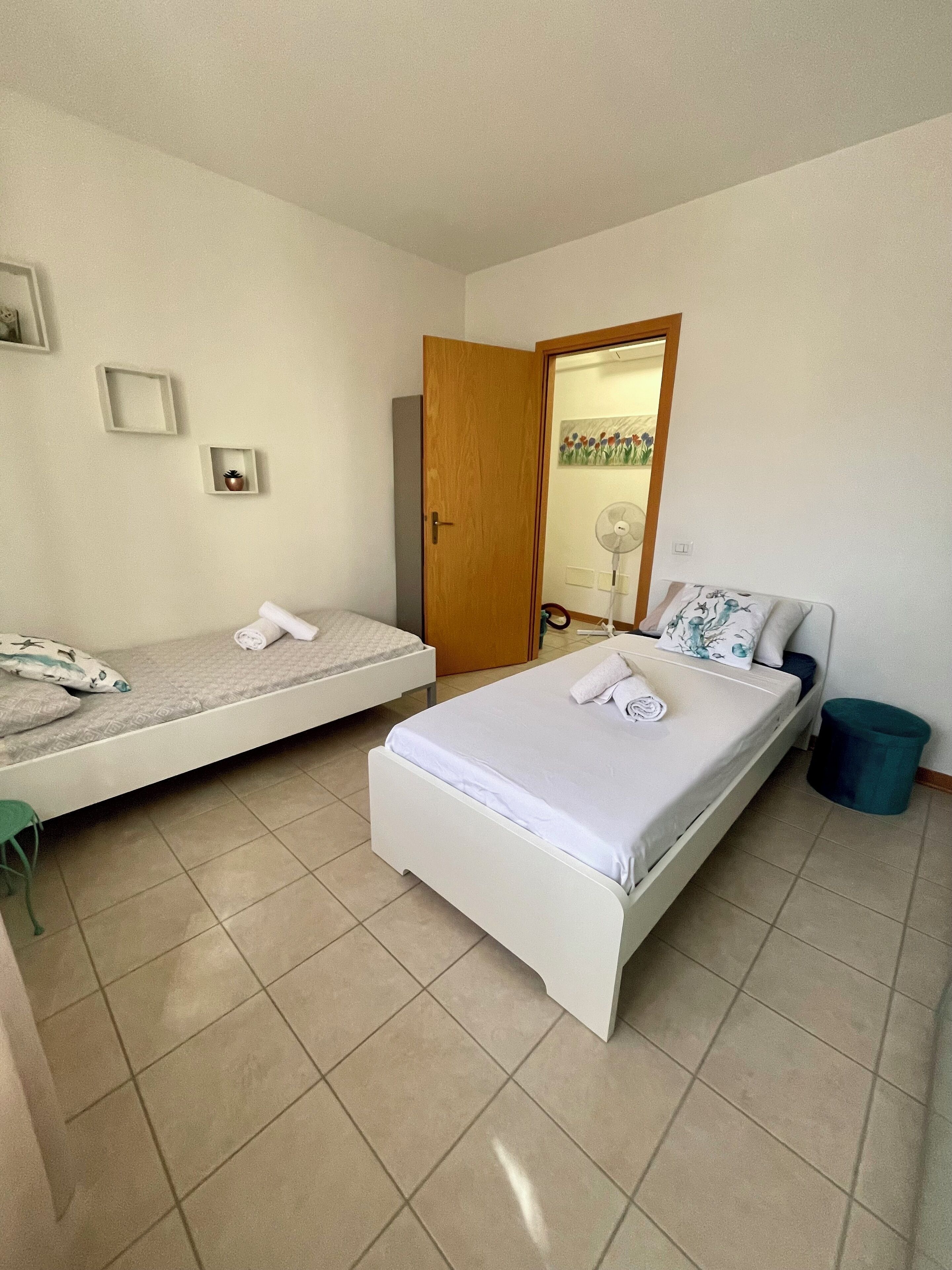 Un dormitorio con dos camas, una puerta, un ventilador y un cubo de basura pequeño.