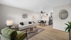 En moderne stue med sofa, sofabord og stole, et spiseområde med bord og stole, og et køkken med skabe og køleskab.