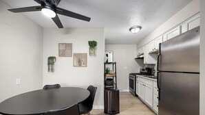 Et moderne køkken med hvide skabe, et rustfrit stål køleskab, et sort spisebord med stole og en loftsvifte.