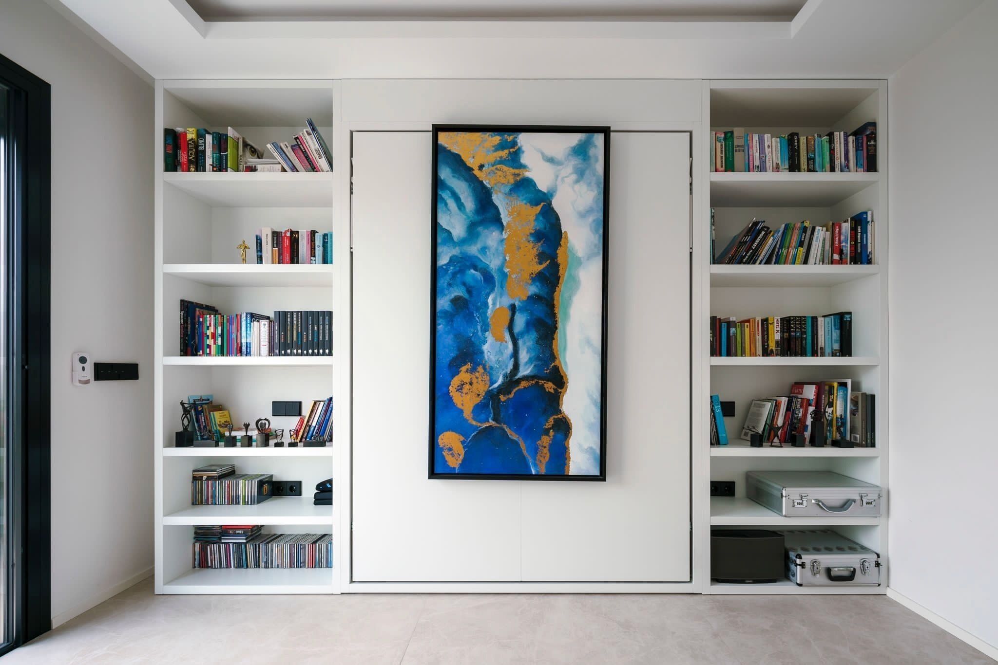 Une pièce aux murs blancs, ornée d’un grand tableau abstrait et de deux étagères blanches remplies de livres et d’objets décoratifs.