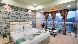 A hotel room with a large bed, a small table, two chairs, a potted plant, and a view of the city through the window.