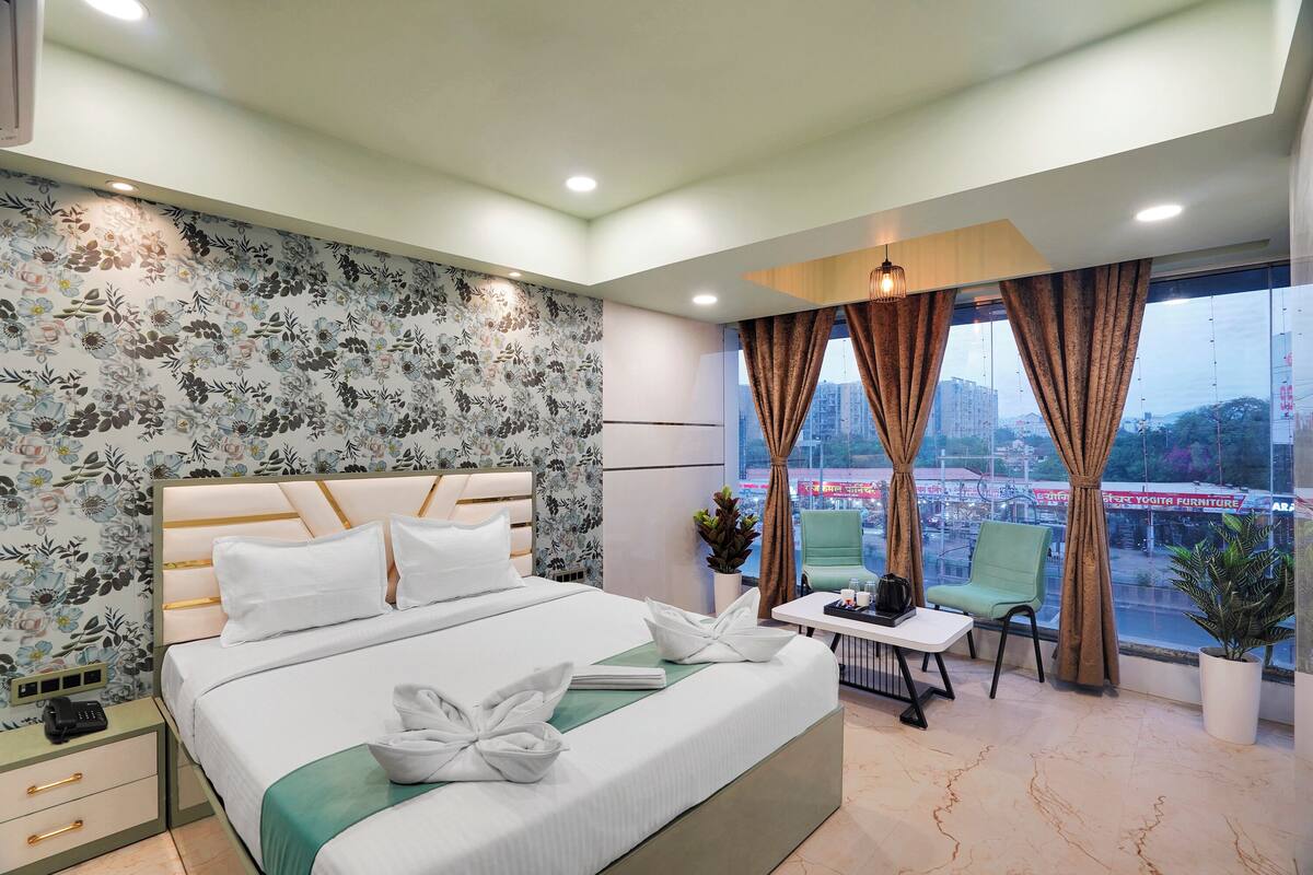 A hotel room with a large bed, a small table, two chairs, a potted plant, and a view of the city through the window.
